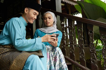 Muslim couple in traditional Malay clothing sitting at wooden stair using smart phone during Eid al-Fitr celebration.の写真素材