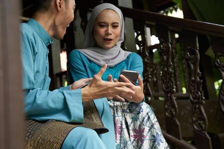 Muslim couple in traditional Malay clothing sitting at wooden stair using smart phone during Eid al-Fitr celebration.の写真素材