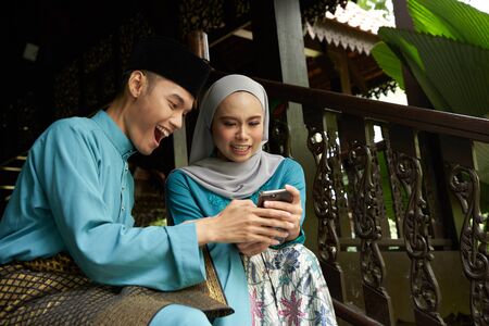 Muslim couple in traditional Malay clothing sitting at wooden stair using smart phone during Eid al-Fitr celebration.の写真素材