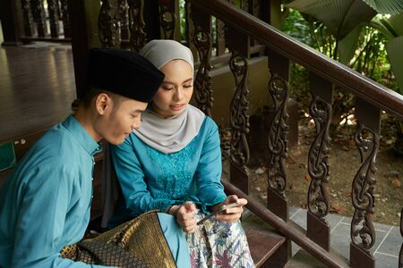 Muslim couple in traditional Malay clothing sitting at wooden stair using smart phone during Eid al-Fitr celebration.の写真素材
