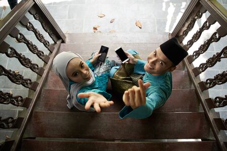 top view of Muslim couple in traditional Malay clothing sitting at wooden stair using smart phone during Eid al-Fitr celebration.