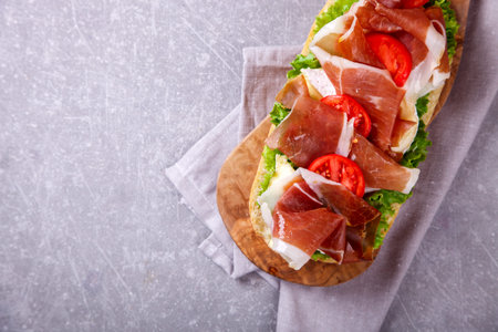 Sandwich with ciabatta, prosciutto,brie cheese and lettuce.on the Board, a grey stone background.の写真素材