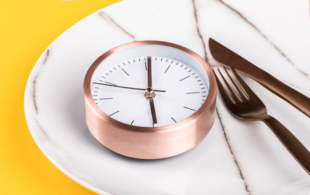 Concept minimal Time to Eat. Alarm Clock on a white Plate on a on yellow Background.Trendy abstract...