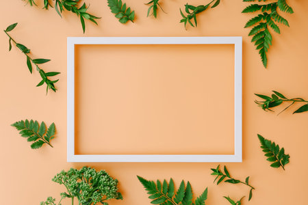 Empty picture frame with green leaves on peach color background, top view