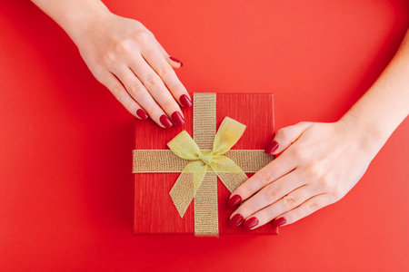 Female hands holding red goft box present with golden bow on red background. Top view
