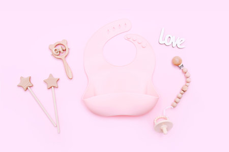 Pink silicone bib and other baby accessories on pink background, top view.