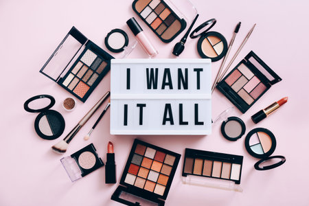 Text I WANT IT ALL on the lightbox. Make up products scattered on pink background, top view