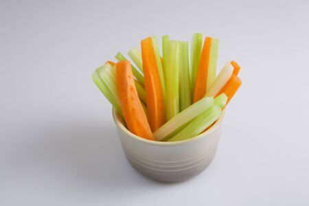 close up filled frame isolated side view shot of a brown beige bowl filled with crunchy orange carrot slices and juicy green celery sticks on a white backgroundの写真素材