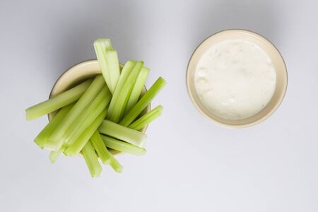 close up isolated flat lay top view shot of a bowl of crunchy juicy green celery sticks next to a white cup of blue cheese dipping sauce on a white backgroundの写真素材
