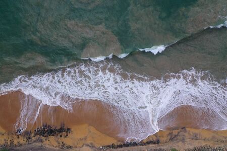 aerial drone bird view shot of the sea shore with yellow sand, black rocks, large white waves and foam crashing on the beach forming beautiful textures, patterns, shapes. Sri Lankaの写真素材