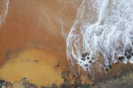 aerial drone bird view shot of the sea shore with yellow sand, black rocks, large white waves and foam crashing on the beach forming beautiful textures, patterns, shapes. Sri Lankaの写真素材