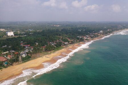 natural aerial drone bird view day shot of the sea shore with a road, beautiful villas, green trees, yellow sand, turquoise blue water, big waves and foam crashing on the beach. Pitiwella, Sri Lankadefaultの写真素材