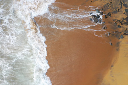 aerial drone bird view shot of the sea shore with yellow sand, black rocks, large white waves and foam crashing on the beach forming beautiful textures, patterns, shapes. Sri Lankaの写真素材