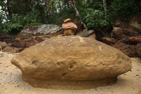 close up shot of a huge brown yellow isolated turtle-shaped rock naturally carved by time by waves with a stack of small stones laying on white sand beach on green jungle forest background. Thailandの写真素材