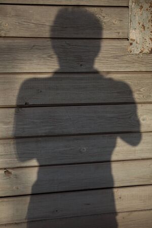 vertical full screen filled frame selfie shot of a silhouette of a man's upper body with sharp contour made by the day sun light on a brown beige wooden plank wallの写真素材