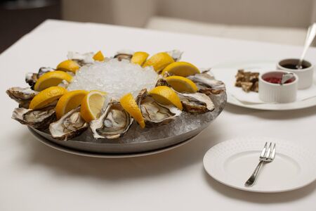 side view close up food shot of fresh raw shucked open oysters lying between lemon slices on a round cold ice tray next to Mignonette shallot vinegar and Ponzu sauces on a white tableの写真素材