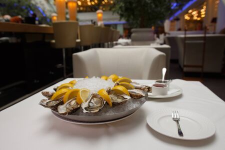 side view close up food shot of fresh raw shucked open oysters lying between lemon slices on a round cold ice tray next to Mignonette shallot vinegar and Ponzu sauces on a white tableの写真素材