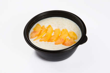 isolated close up top corner view shot of a delicious white rice milk porridge with peach fruit slices on top in a black plastic bowl on a white backgroundの写真素材