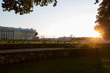 SAINT PETERSBURG, RUSSIA 08/13/2021 people sitting resting on the green grass of a field of Alexander Garden Park. Bronze Horseman sculpture, golden hour summer sunset sun, blue sky in the background.のeditorial素材