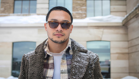 A serious young asian man in brown faux fur coat and sunglass standing outdoors in winter snow. confident, stylish portrait in cold weatherの素材
