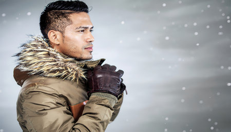 A handsome young asian man feels cold in winter parka with fur hood, wearing gloves and looking away during snowfall with pensive lookの素材