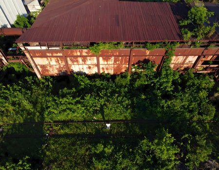 Desolate aerial view of an abandoned, rusty, and overgrown industrial factory. Nature reclaims desolate and decaying building with green vine, creating an eerie post apocalyptic sceneの素材