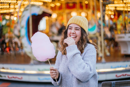 Young girl holding a cotton candy and smiling at a Christmas fair wearing a yellow wool cap and a...