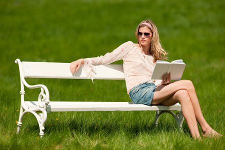 Young blond woman sitting on white bench with book; shallow DOFの写真素材