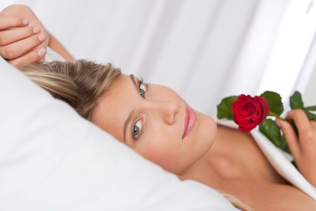 Beautiful woman lying in white bed holding red roseの写真素材