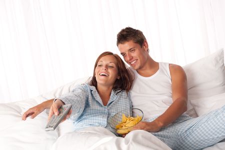 Young man and woman lying down in white bed watching television and eating crispsの写真素材