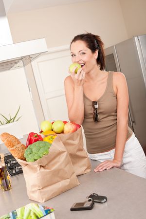 Young smiling woman with groceries in the kitchen biting appleの写真素材