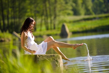 Happy romantic woman sitting by lake splashing water, wearing white dressの写真素材