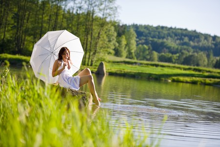 Happy romantic woman sitting by lake with parasol on sunny dayの写真素材