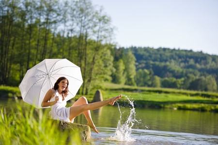 Happy romantic woman sitting by lake splashing water, holding parasol, wearing white dressの写真素材