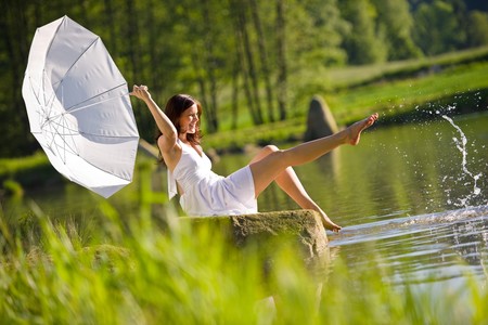 Happy romantic woman sitting by lake splashing water, holding parasol, wearing white dressの写真素材