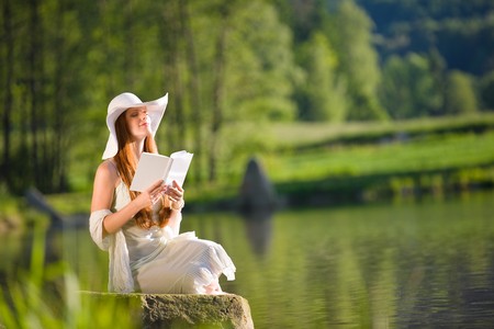 Long red hair romantic woman relax by lake with book on sunny dayの写真素材