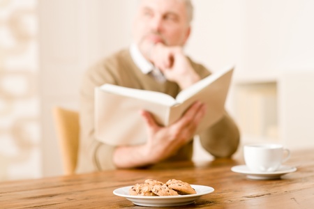 Senior mature man reading book, cookies at wooden tableの写真素材