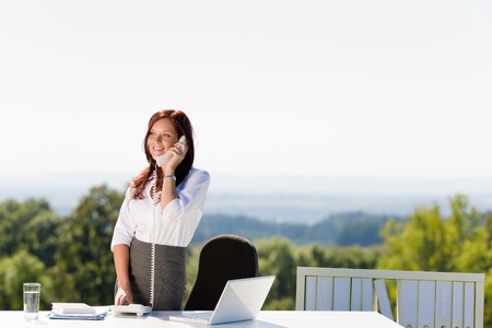 Young businesswoman in sunny nature office calling standing behind tableの写真素材