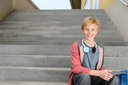 Happy teenage student boy with tablet sitting on steps outdoorsの写真素材