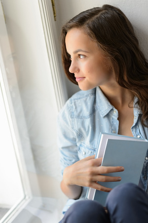 Beautiful teenage girl student holding book looking out the windowの写真素材
