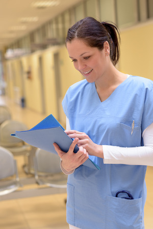 Smiling young female doctor in hospital looking down at patient records standing alone in hallwayの写真素材