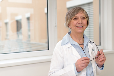 Senior doctor standing in hospital smiling holding her glassesの写真素材