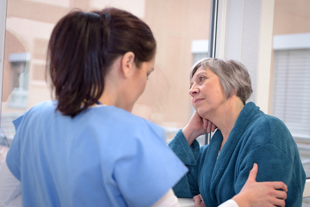 Sad senior patient in hospital corridor looking outside with young nurseの写真素材