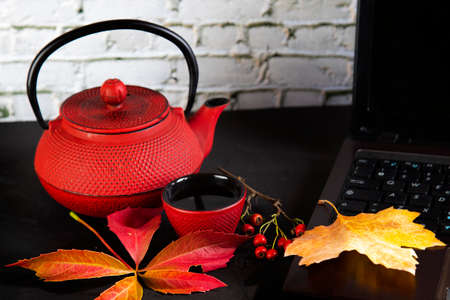 on a black table a laptop, a red teapot with tea, a red cup and autumn leaves and berriesの写真素材