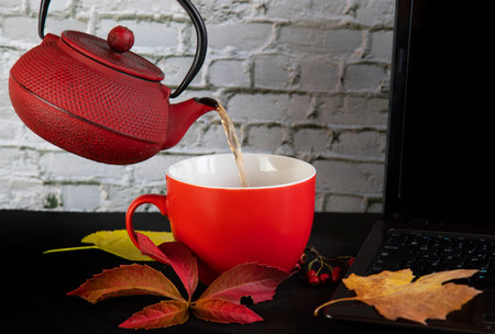 on a black table a laptop, a red teapot with tea, a red cup and autumn leaves and berriesの写真素材