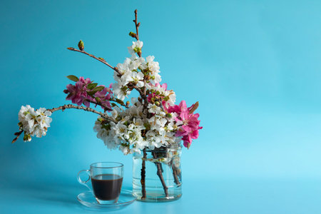 transparent cup of coffee next to a glass vase with flowering spring branches on a blue background