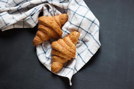 Two fresh croissant lying on a blackboard. Also, there is twisted linen towel. Croissants are delicious and appetizing. Towel is white with blue pattern.の写真素材