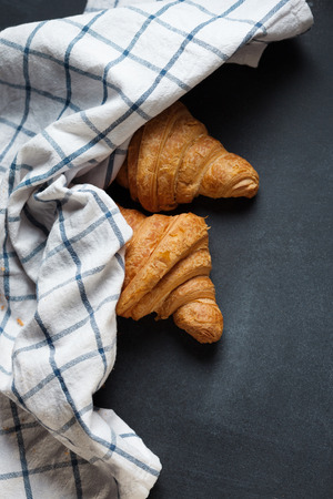 Two fresh croissant lying on a blackboard. Also, there is twisted linen towel. Croissants are delicious and appetizing. Towel is white with blue pattern.の写真素材