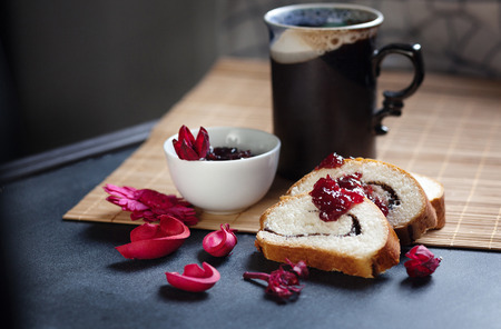 Slices of butter poppy roll, served with cherry jam and large ceramic cup with hot drink. Black table and bamboo mat. Red dry flowers are scattered around. Ceramic mug has a beautiful decorative handle.の写真素材