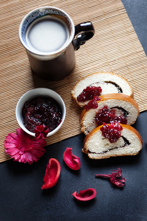 Slices of butter poppy roll, served with cherry jam and large ceramic cup with hot drink. Black table and bamboo mat. Red dry flowers are scattered around. Ceramic mug has a beautiful decorative handle.の写真素材
